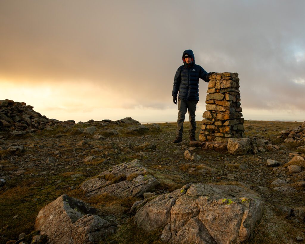 Simon Evans tapping the trig point at the summit of Pillar during a winter sunset in the Lake District.
