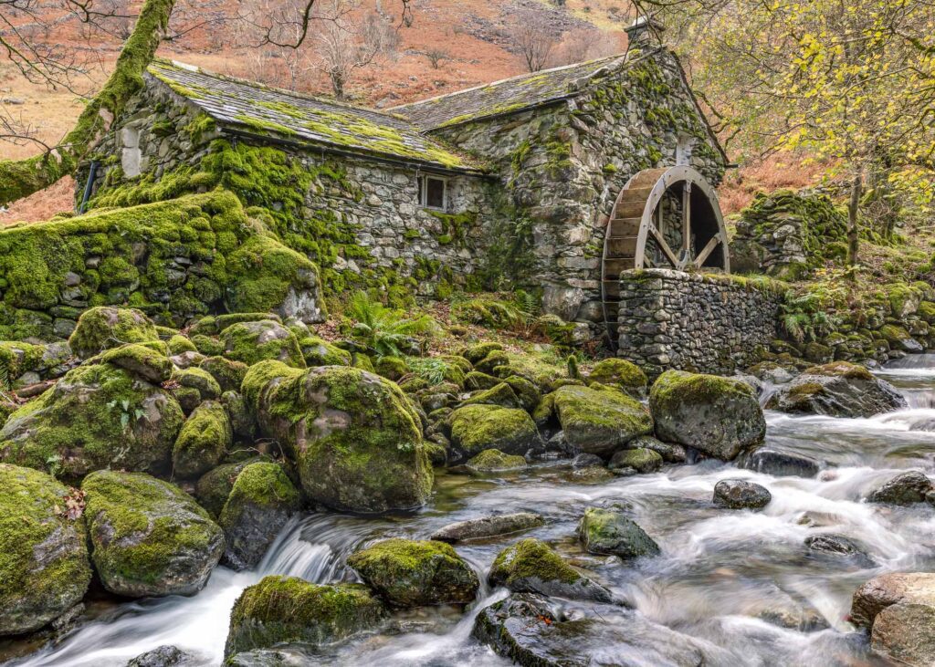 Lake District landscape photography. In the heart of the English Lake District stands a hidden watermill, perched on the bank of a fast-flowing river. In this photograph, the old stone mill dominates the frame with the roaring river streaking through the foreground. The mountain in the distance behind the mill provides a beautiful autumnal backdrop for the frame. This photograph was captured with a Canon 24-70mm F2.8L lens.