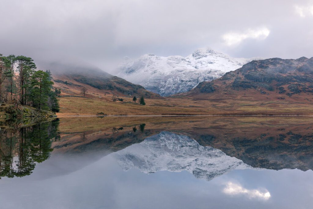 Lake District landscape photography. This image is captured from the beautiful iconic shores of Blea Tarn during the depths of winter. Recent snowfall blankets the Langdale Pikes dominating the background with its spectacular reflection projected across the perfectly calm tarn. This photograph was captured with a Canon 24-70mm F2.8L lens.