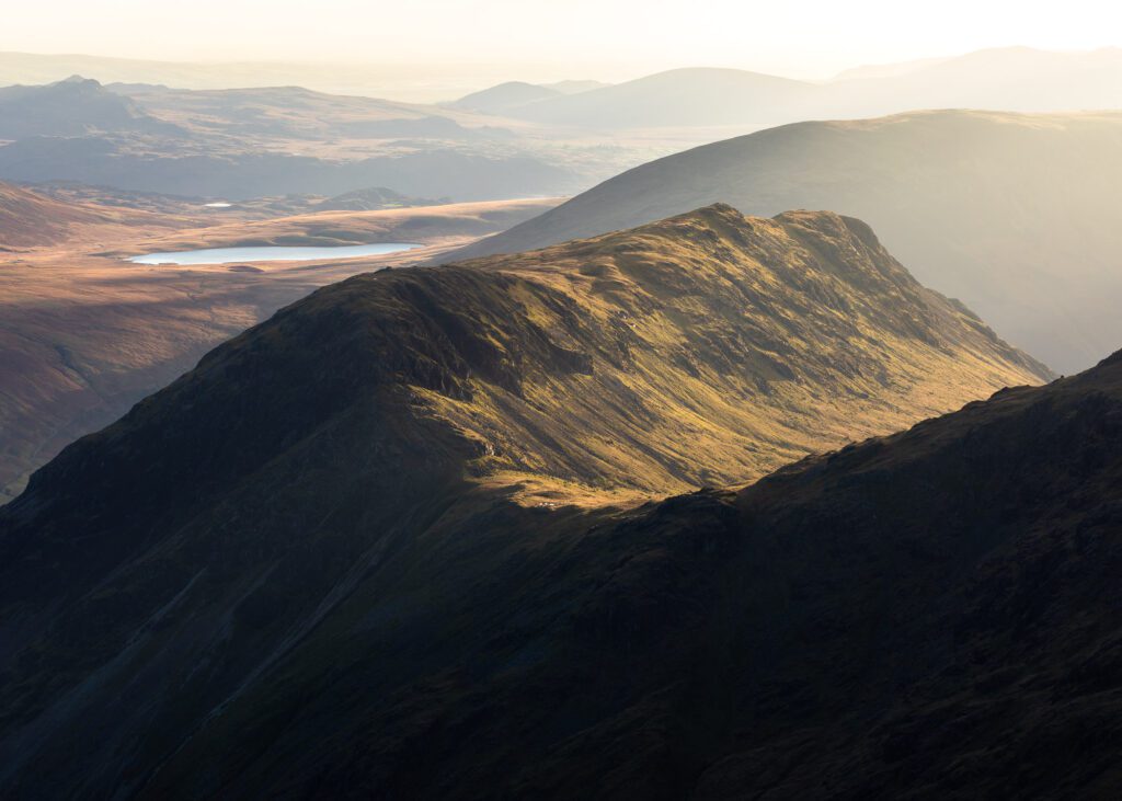 Lake District landscape photography. The beautiful curves of Yewbarrow's ridge guide you through this image. In the distance, Illgill Head and Burnmoor Tarn stand draped in stunning golden hour light with the distance fells shrouded with a blanket of haze. This photograph was captured with a Canon 70-200mm F4L lens.