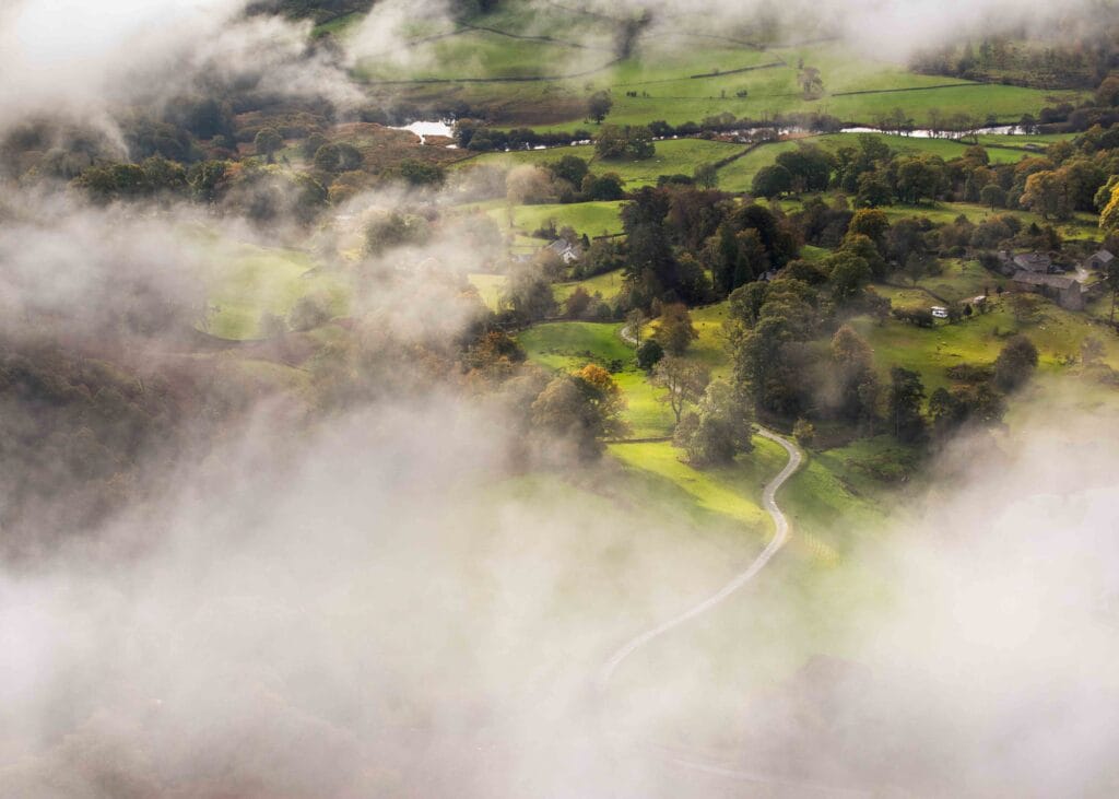 A breathtaking temperature inversion begins to lift as the sun rises over Loughrigg Fell revealing Loughrigg Tarn and Fell Foot Farm in the Lake District.