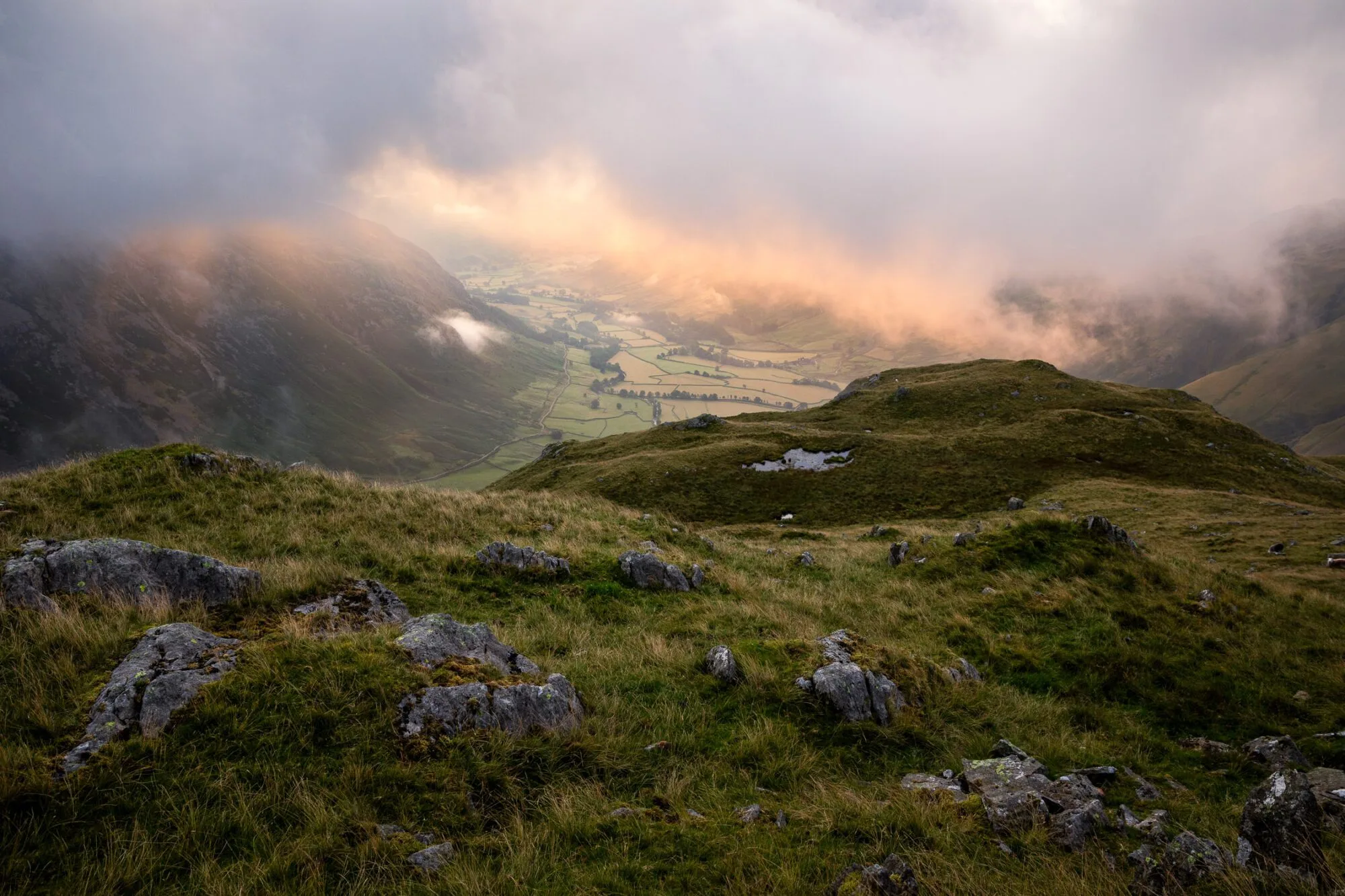 A view from the band on the ascent of Bowfell in the Lake District during a breathtaking summer morning. Low cloud hovers above Mickleden Valley catching the glorious sunrise light around Great Langdale.