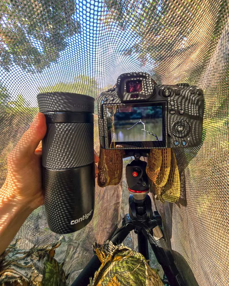 Nature photographer Simon Evans enjoying a vacuum mug of tea under a makeshift camouflage hide on a lake, waiting for Kingfishers to show.