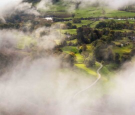A breathtaking temperature inversion begins to lift as the sun rises over Loughrigg Fell revealing Loughrigg Tarn and Fell Foot Farm in the Lake District.